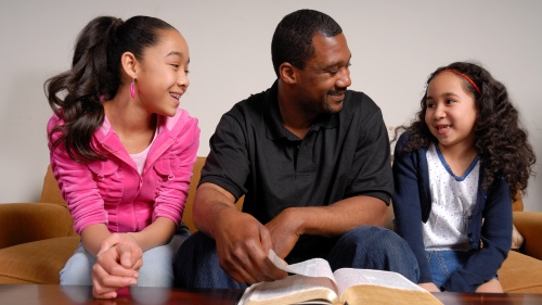 A dad and his two daughters talking with a Bible opened.